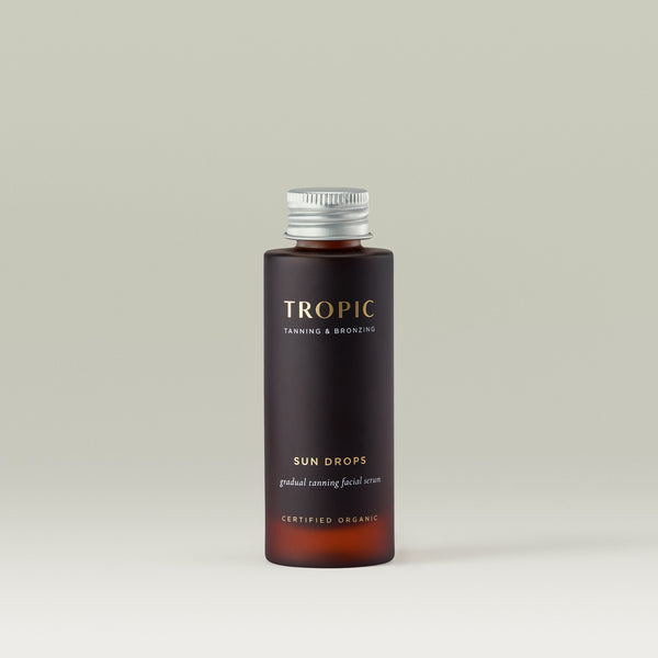 Tropic Sun Drops gradual tanning facial serum refill bottle in amber glass with a silver screw cap, photographed against a soft neutral background. The label features gold and white text with 