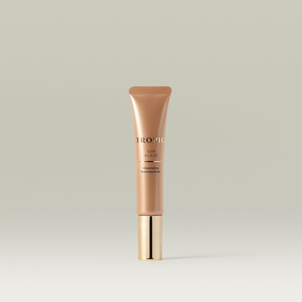 A sleek bronze tube of Tropic Sun Glaze illuminating bronzing drops with a shiny gold cap, standing upright against a soft neutral background.