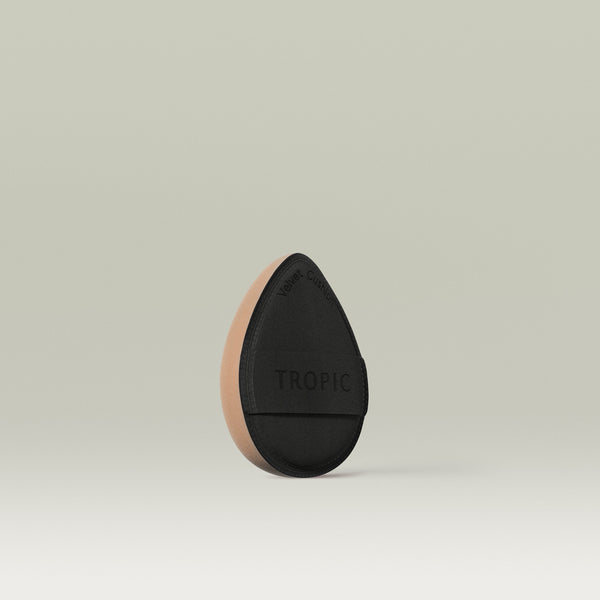 A teardrop-shaped Tropic velvet makeup cushion with a black textured surface, a finger strap across the front, and a soft beige underside, shown standing upright against a neutral background.