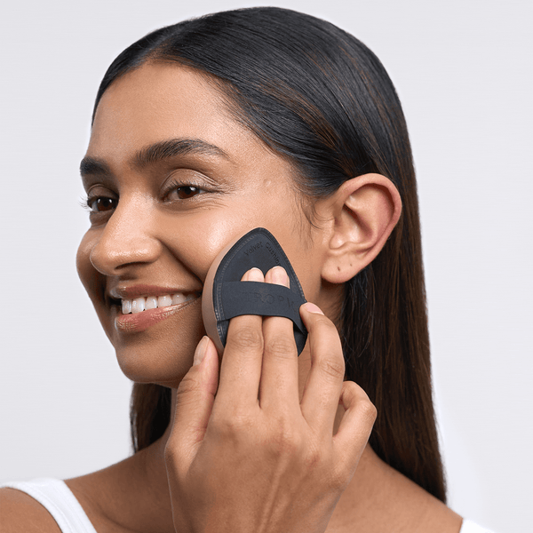 A smiling woman applies makeup using a black, teardrop-shaped blending sponge with a finger strap, gently pressing it against her cheek.